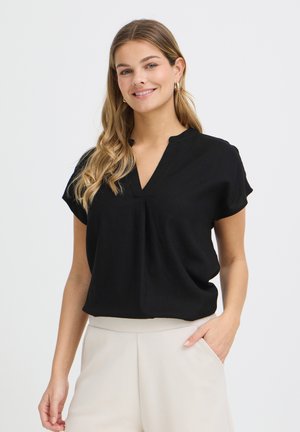 Lächelnde Frau mit langen, welligen Haaren, die eine schwarze V-Neck-Bluse mit kurzen Ärmeln und beige Hosen trägt, mit einer Hand in der Tasche.