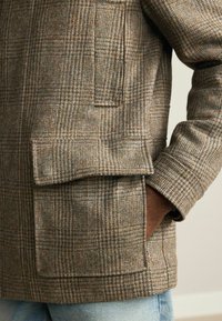 Pruunikirjuuline mantel, mis on valmistatud tekstureeritud villasegistist, millel on kahekordne põhipocket ja mansetiga varrukas. Mustril on peened sinised aktsendid.