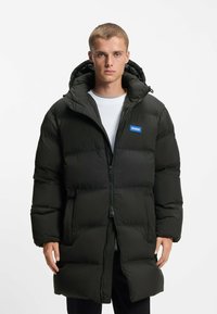 Vaatteena musta pehmustettu parka-takki hupulla, etuvetoketjulla ja sivutaskuilla; sisältää sinisen logopatchin. Sujuva rakenne, ylisuurikokoinen muoto.