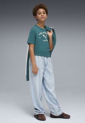 Camiseta de manga corta color verde azulado con logo blanco, vaqueros sueltos azul claro y zapatos marrones. Suéter con capucha colgado sobre un hombro.