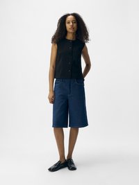 Top negro sin mangas con botones en la parte frontal, combinado con pantalones cortos tipo bermuda de mezclilla en un tono azul medio. La modelo lleva mocasines negros. Diseño minimalista.