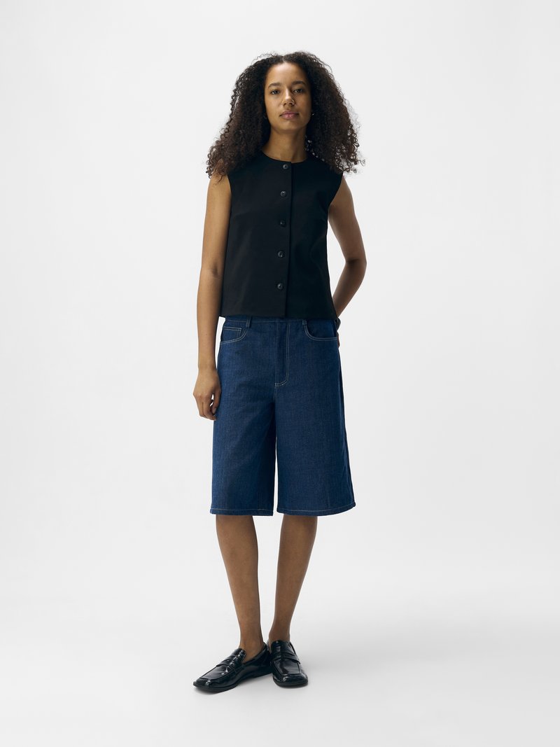 Top negro sin mangas con botones en la parte frontal, combinado con pantalones cortos tipo bermuda de mezclilla en un tono azul medio. La modelo lleva mocasines negros. Diseño minimalista.