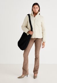 Lyse beige quiltet jakke med høj krave, brune cordbukser og en struktureret sort taske. Har spidse beige hæle med en blank finish.