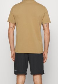 Béžové polo tričko s krátkými rukávy, rovný spodní lem a klasický límec, spojené s černými šortkami, které mají malý logo na noze.