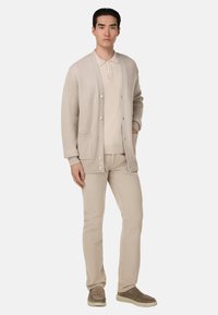 Beżowy kardigan o prążkowanej fakturze, z pięcioma guzikami i dwiema kieszeniami; zestawiony z jasnobeżową koszulą bez kołnierzyka i pasującymi spodniami.