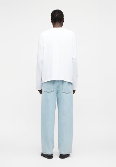 Άτομο που στέκεται με την πλάτη προς τα εμάς, φορώντας μια χαλαρή λευκή μπλούζα με μακριά μανίκια, ανοιχτού μπλε φαρδιά τζιν και μαύρα παπούτσια σε απλό φόντο.