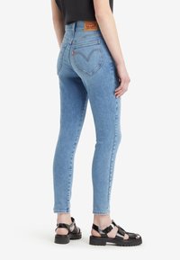 Jeans de corte ajustado en azul claro con una cintura alta, presentan un parche de cuero en la parte posterior y bolsillos traseros en ángulo. Combinados con sandalias negras de tiras.