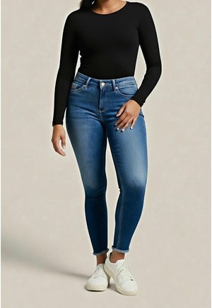 Kvinde iført en sort langærmet top, blå højtaljede skinny jeans og hvide sneakers, står mod en ensfarvet baggrund.