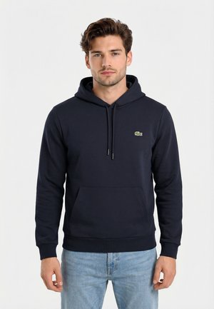 Giovane uomo con capelli castani che indossa una felpa con cappuccio blu scuro e jeans azzurri, in piedi contro uno sfondo chiaro e uniforme.