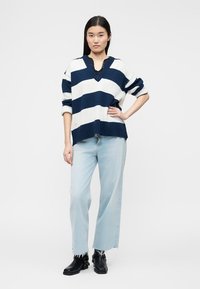 Γυναίκα που στέκεται με το χέρι στη μέση, φοράει πουλόβερ με ναυτικό και λευκό ριγέ, ανοιχτόχρωμο τζιν με ξεφτισμένα τελειώματα και μαύρα μποτάκια αστραγάλου.