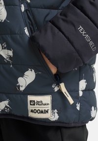 Blå vadderad väst med Mumin-mönster, med en dragkedja fram, logotypmärke och TEXASHIELD-etikett på ett mörkt tyg. Mjuk textur, lättviktig design.