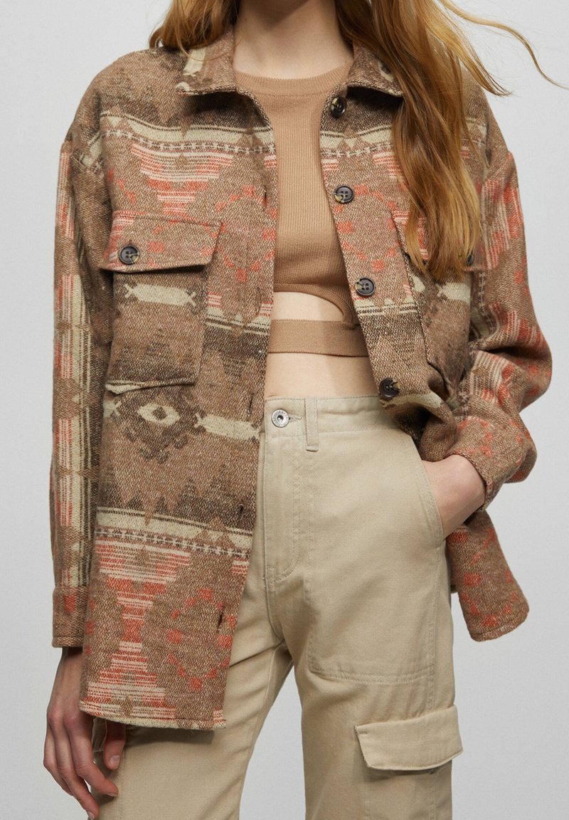 Femme portant une veste marron et beige à motifs sur un haut court beige, associée à un pantalon cargo beige taille haute, main dans la poche.