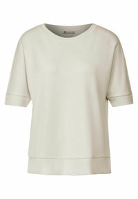 Světlý béžový krátký rukáv s kulatým výstřihem a volným střihem. Má hladkou texturu a jednoduché šicí detaily podél spodního okraje.