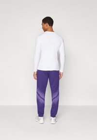 Camisola de manga longa branca combinada com calças de treino roxas com efeito tie-dye, apresentando um cós elástico e um corte relaxado. Footwear são sapatilhas desportivas de tom claro.