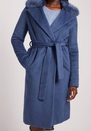 Frau trägt einen blauen Wollmantel mit Gürtel und Fellkragen und einen hellblauen Strickpullover darunter, steht mit einer Hand in der Tasche.