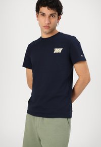 T-shirt en coton bleu marine à manches courtes, avec un imprimé crème sur la poitrine gauche et une petite étiquette sur la manche.