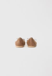 Scarpe basse in suede marrone con una forma arrotondata, caratterizzate da un sottile accento beige nella parte superiore e da lacci sottili per la chiusura. Suola liscia.