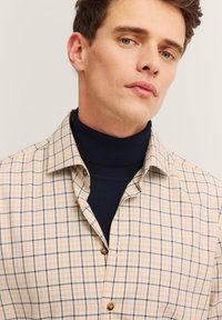 Camicia a quadri beige con colletto strutturato, caratterizzata da controlli blu e arancioni, abbinata a un dolcevita navy scuro sotto.