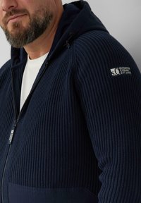 Brodaty mężczyzna noszący granatowy, teksturowany hoodie z zamkiem, z białą koszulą w środku i naszywką "Elevated Casual Est. 1969" na rękawie.