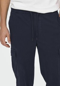 Marine Cargo-Hosen mit einem elastischen Bund, Kordelzug, seitlichen Taschen und einer aufgesetzten Tasche am Oberschenkel, die eine glatte Textur aufweisen.