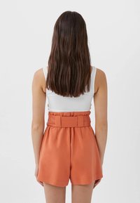 Shorts taille haute orange avec une ceinture, fabriqués dans un tissu lisse. Un design uni avec des plis et une coupe décontractée. Débardeur blanc visible.