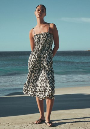 Frau steht am Strand, trägt ein schwarz-weiß gemustertes Midikleid mit dünnen Trägern und schwarze Sandalen unter klarem blauem Himmel.