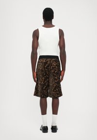 Homme de dos portant un débardeur blanc, un short à motifs marron et noir arrivant au genou, des chaussettes blanches et des chaussures noires sur fond uni.