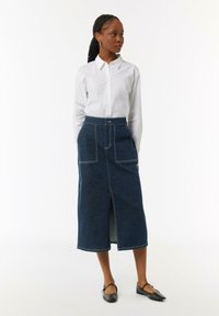 Dunkelblaue Jeansrock mit vorderem Schlitz, kontrastierendem weißen Nähten und zwei großen Taschen. Kombiniert mit einem weißen Hemd und schwarzen Ballerinas.