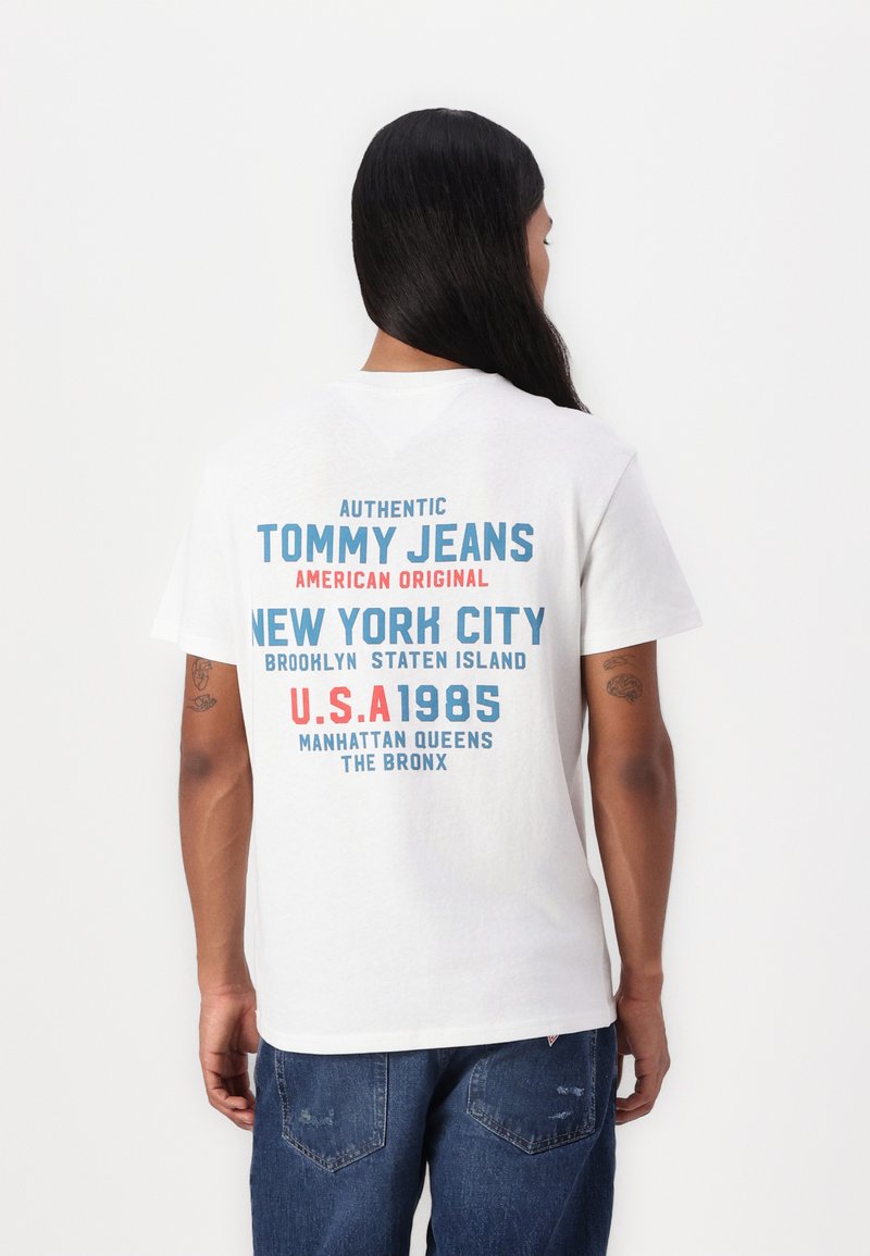 T-shirt en coton blanc avec un texte multicolore au dos : "AUTENTIQUE TOMMY JEANS AMÉRICAIN ORIGINAL," avec des graphiques de ville et d'année.