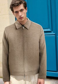 Braune Wolljacke mit Kragen, Reißverschluss vorne und Seitentaschen. Verfügt über einen strukturierten Stoff und einen gerippten Saum. Wird über einer hellen Hose getragen.