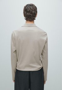 Beige poloshirt met lange mouwen en kraag, met een gladde structuur en een ontspannen pasvorm, gecombineerd met een donkere broek. Achteraanzicht.