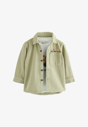 Béžová košeľa na gombíky s náprsným vreckom, vyšívané hračkárske autíčka nad vreckom, nosená na bielom tričku s potlačou.