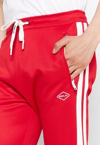 Rote Sporthose mit weißen Seitenstreifen, ausgestattet mit einem Kordelzugbund und einer Reißverschlusstasche. Glatter Stoff und Rauten-Logo-Detail.