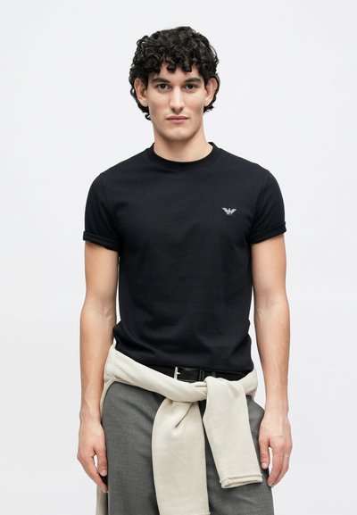 Juoda medvilninė marškinėliai su apvalia iškirpte ir surolleintomis rankovėmis, priekyje mažas erelio logotipas. Laisvo stiliaus, dėvimi su pilkomis kelnėmis.