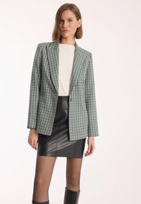 Zielony i fioletowy żakiet w wzór houndstooth z jednym guzikiem, w zestawieniu z czarną skórzaną mini spódnicą i czarnymi botkami do kolan.