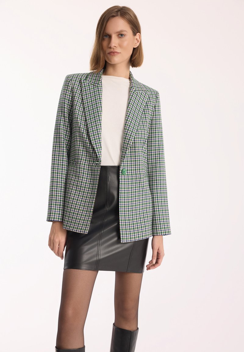 Zielony i fioletowy żakiet w wzór houndstooth z jednym guzikiem, w zestawieniu z czarną skórzaną mini spódnicą i czarnymi botkami do kolan.