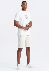 Hvid bomulds t-shirt med et cirkulært grafisk design; matchende med lysebeige shorts og hvide sneakers, har en afslappet pasform og lommer.