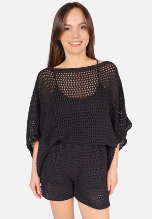 Jeune femme portant un haut en maille noire ajourée et un short assorti, souriante, avec de longs cheveux bruns et raides sur un fond uni.
