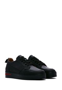 Baskets en cuir noir avec un bout rond, semelle surélevée, lacets noirs et une bande rouge contrastante sur la semelle. Texture lisse et design minimal.