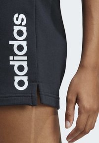 Čierne športové šortky s bielym logom "adidas". Majú bočné rozparky na leme a rebrovanú textúru. Detailný záber ruky na šortkách.