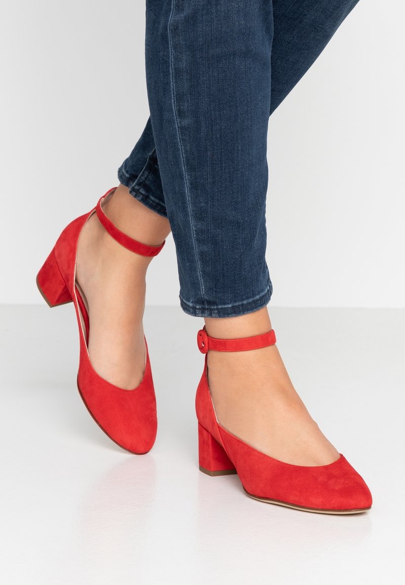 Pieds portant des chaussures rouges en daim à talon bloc avec bride à la cheville, associées à un jean bleu foncé retroussé sur un sol blanc.