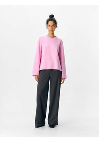Maglietta rosa a maniche lunghe con scollatura rotonda, abbinata a pantaloni larghi grigio scuro. Entrambi i capi hanno una texture liscia e uno stile casual.