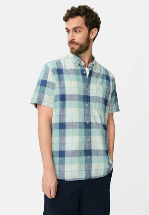 Homme portant une chemise à manches courtes à boutons avec un motif à carreaux bleu et vert, debout avec une main dans la poche, regardant sur le côté.