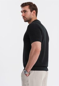 Suéter de punto negro de manga corta con dobladillo y cuello acanalados, combinado con pantalones de color claro. La modelo tiene las manos en los bolsillos.