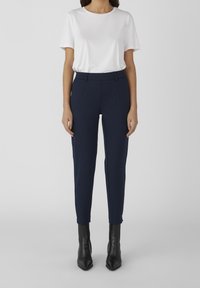 Bílý tričko s krátkým rukávem spárované s tmavě modrými zúženými kalhotami a černými kotníkovými botami, s hladkou texturou a přizpůsobeným střihem.