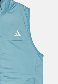 Ljusblå ärmlös väst med kviltad textur, med en liten vit triangulär logotyp och en slät, rund kragdesign.