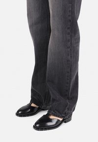 Schwarze Leder-Slipper mit offenen Zehen, flachem Absatz und silbernen Nietenakzenten an der Sohle, kombiniert mit grauen geraden Jeans.