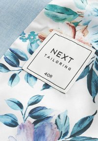 Květinová látka s modrými a fialovými listy a květinami, na modrém strukturovaném pozadí. Štítek zní "NEXT TAILORING 40R."