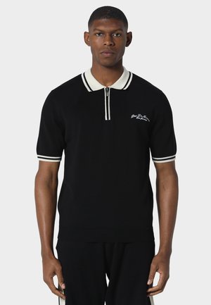 Mand stående vendt fremad iført sort kortærmet poloshirt med hvid krave, manchetter og lille broderet tekst på brystet.