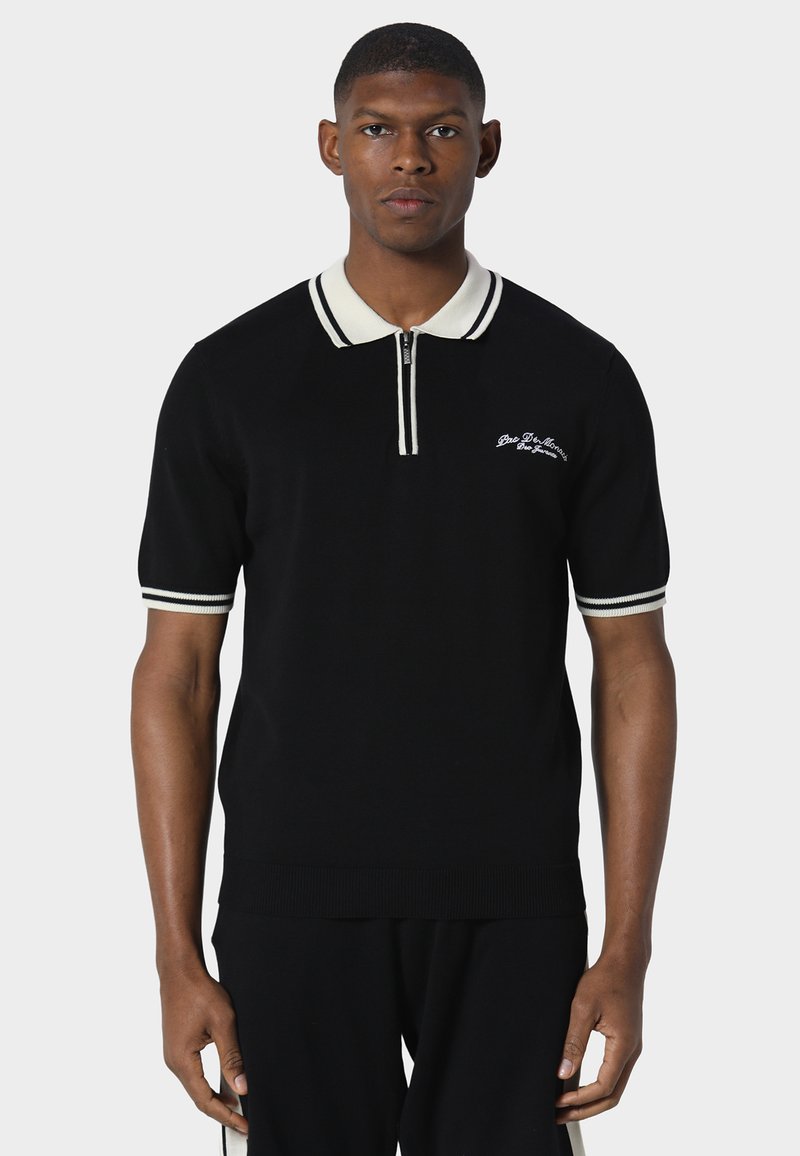 Homme debout face à l'avant portant un polo noir à manches courtes avec col blanc, poignets blancs et un petit texte brodé sur la poitrine.